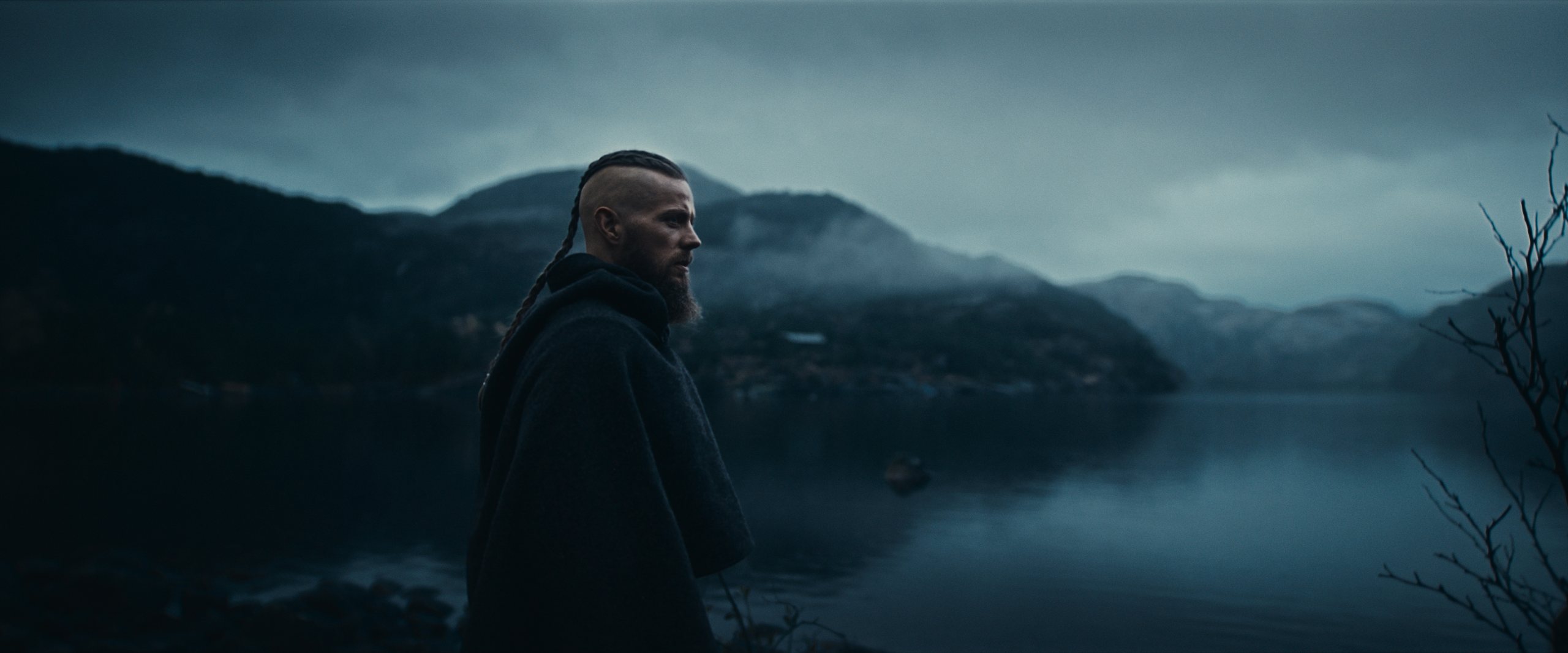 A man in a dark wool cloak on the shore of a misty Nordic fjord at dusk.